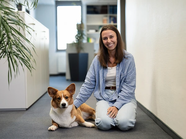 Eine Frau sitzt im Büro auf dem Boden und streichelt einen Hund, der neben ihr liegt. Im Hintergrund sind Regale und Pflanzen sichtbar.