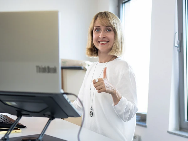 Eine Frau steht lächelnd auf einem Laufband in einem hellen Büro, während sie an einem Computer arbeitet und Daumen nach oben zeigt.
