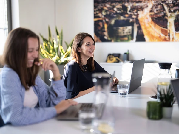 Zwei Frauen sitzen an einem Tisch in einem modern eingerichteten Büroraum; eine Frau lächelt, während die andere lachend auf ihrem Laptop arbeitet. Im Hintergrund ist ein großes Bild mit einer Stadtansicht zu sehen. Auf dem Tisch stehen Wassergläser und Pflanzen.