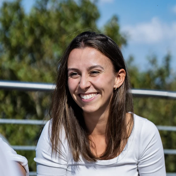 Eine fröhliche Frau mit langen, braunen Haaren sitzt an einem Tisch im Freien und lächelt, während sie mit anderen Menschen spricht. Der Hintergrund zeigt grüne Bäume und einen blauen Himmel mit einigen Wolken.