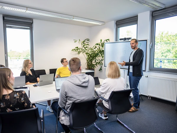 Eine Gruppe von sieben Personen sitzt an einem Konferenztisch in einem hellen Meetingraum. Der Vortragende, ein Mann, steht vor einem Whiteboard und präsentiert während einer Besprechung. Alle Teilnehmer nutzen Laptops und sind aufmerksam. Im Hintergrund ist eine große Pflanze und Fenster mit Tageslicht zu sehen.