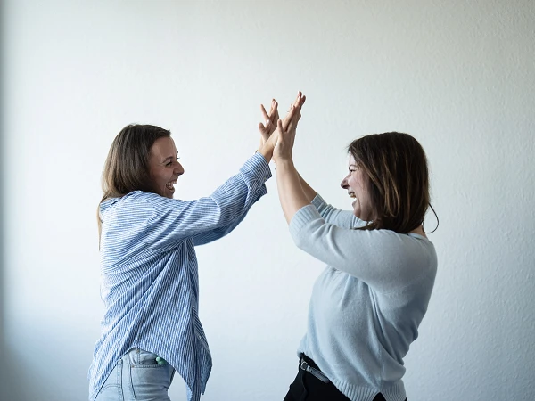 Zwei Frauen stehen sich gegenüber und klatschen einander in die Hände, während sie lachen und offensichtlich Spaß haben. Der Hintergrund ist eine helle Wand und ein Fenster, das natürliches Licht hereinlässt.
