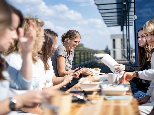 Eine Gruppe von Frauen sitzt an einem langen Tisch im Freien und interagiert freundlich miteinander, während sie an verschiedenen Projekten oder Aktivitäten arbeiten. Die Sonne scheint, und im Hintergrund sind moderne Gebäude und ein blauer Himmel zu sehen.