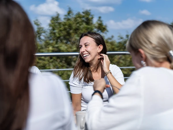 Eine lächelnde Frau mit langen braunen Haaren sitzt an einem Tisch im Freien, umgeben von Personen, die teilweise im Bild sichtbar sind, während sie in einem entspannten Gespräch vertieft sind. Im Hintergrund ist ein blauer Himmel mit einigen Wolken und grüne Bäume zu sehen.