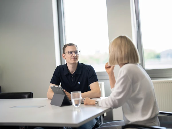 Ein Mann in einem schwarzen Poloshirt sitzt an einem Tisch und schaut eine Frau an, die ihm gegenüber sitzt. Die Frau trägt ein weißes Oberteil und spricht. Auf dem Tisch steht ein Glas Wasser und ein Tablet. Im Hintergrund sind Fenster mit Blick auf eine städtische Landschaft.