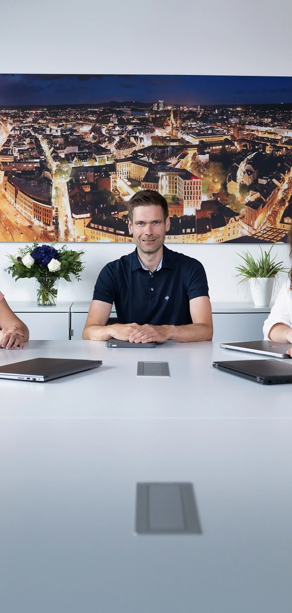 Fünf Personen sitzen an einem großen Tisch in einem modernen Konferenzraum, umgeben von Pflanzen und mit einer Stadtansicht im Hintergrund. Sie schauen lächelnd in die Kamera.