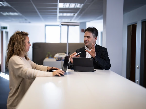 Ein Mann und eine Frau stehen an einem Stehtisch in einem modernen Büro und führen ein Gespräch, während der Mann ein Tablet in der Hand hält und der Frau aufmerksam zuhört.