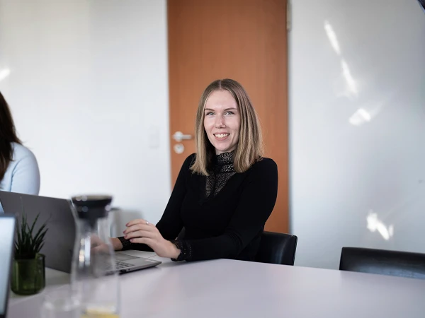 Zwei Frauen sitzen an einem Tisch in einem hellen Büroraum, während die Frau rechts lächelnd in die Kamera schaut und an einem Laptop arbeitet. Auf dem Tisch steht eine Pflanzen dekorative und ein Glaskaraffe.