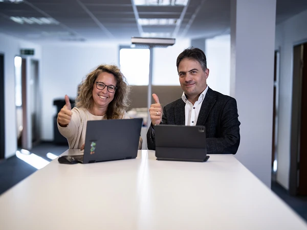 Eine Frau und ein Mann sitzen an einem Tisch in einem Büro, beide zeigen mit Daumen nach oben, während sie auf ihre Laptops schauen.
