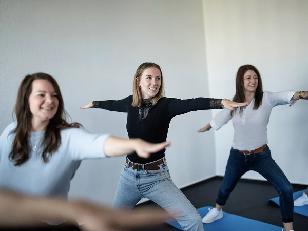 Drei Frauen führen gemeinsam eine Yoga-Übung in einer hellen, modernen Umgebung durch, wobei sie in einer Krieger-Pose stehen und ihre Arme zur Seite ausstrecken.