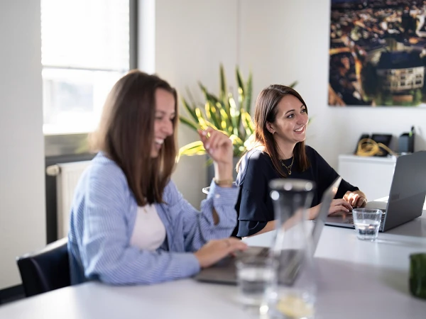 Zwei Frauen sitzen an einem Tisch, lächeln und arbeiten an ihren Laptops in einem hellen Büro mit Pflanzen im Hintergrund.