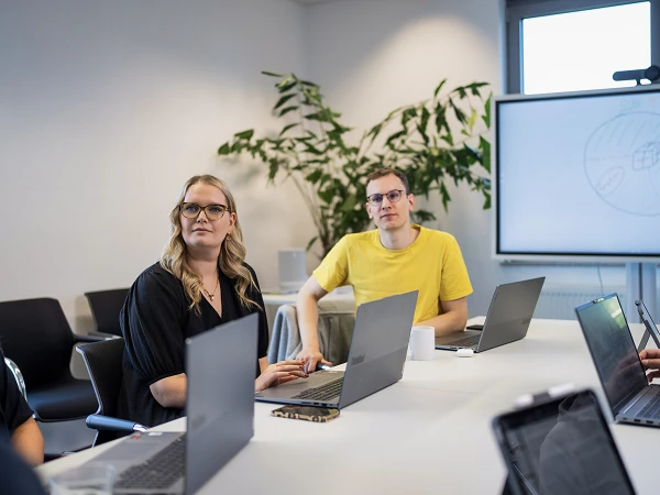 Eine Gruppe von Personen sitzt um einen Tisch in einem Konferenzraum, während sie an Laptops arbeiten und einem Bildschirm mit einer Präsentation im Hintergrund folgen. Zwei der Personen, eine Frau mit langen Haaren und Brille sowie ein Mann mit kurzen Haaren in einem gelben T-Shirt, schauen aufmerksam in die Richtung des Sprechers. In der Ecke des Raumes steht eine große Pflanze.