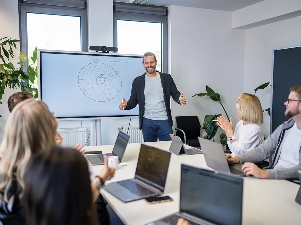 Ein Präsentator steht vor einer Gruppe von fünf Personen in einem Besprechungsraum und zeigt auf eine Tafel mit einer grafischen Darstellung. Die Zuhörer sitzen an einem Tisch mit Laptops und applaudieren, während der Präsentator lächelt und Daumen hoch zeigt. Im Hintergrund sind Pflanzen und Fenster sichtbar.