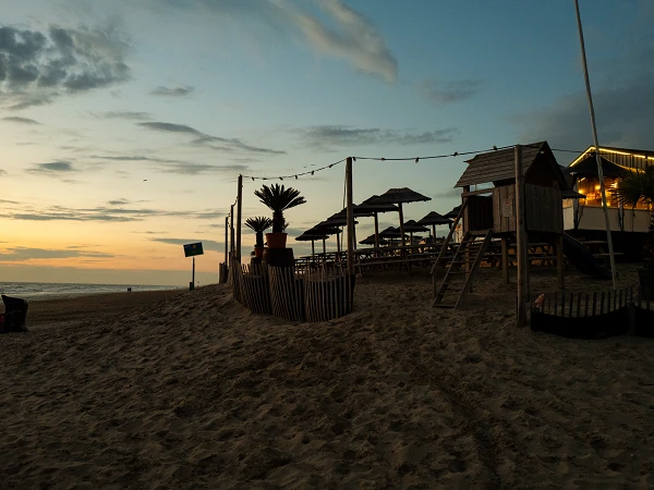 Eine Strandlandschaft bei Sonnenuntergang, mit einer Spielstruktur aus Holz, Liegestühlen unter Sonnenschirmen und einem gemütlichen Restaurant im Hintergrund, umgeben von sandigem Gelände und spärlicher Beleuchtung.