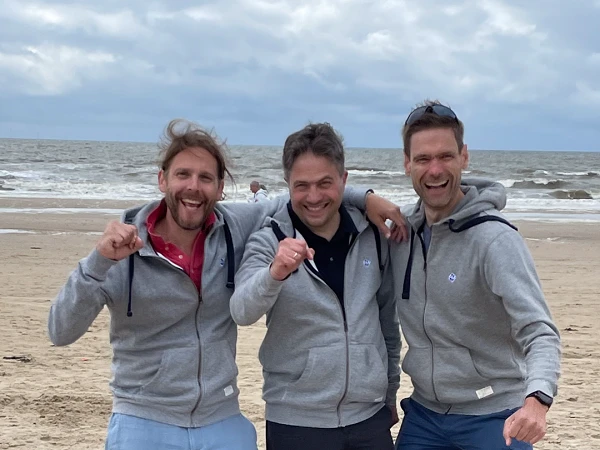 Drei Männer in grauen Kapuzenpullovern stehen am Strand, lächeln und zeigen Daumen hoch, während sie vor dem Meer pic.twitter.com. Der Himmel ist bewölkt und der Sandstrand ist gut sichtbar.