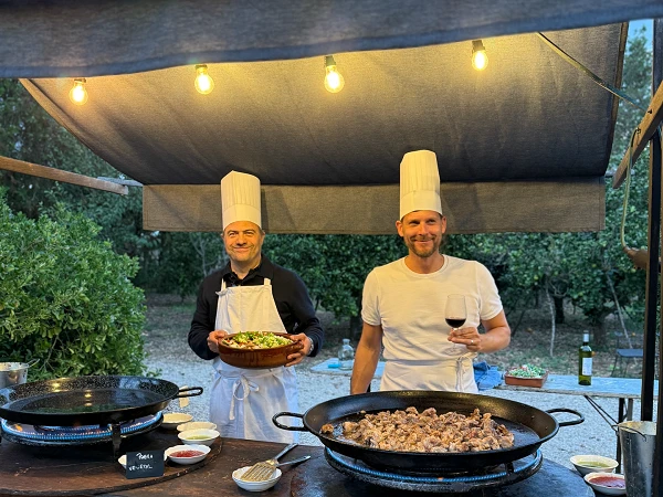 Zwei Köche in Kochmützen stehen an einem Freiluftküchenstand, der mit drei großen Pfannen mit Essen ausgestattet ist. Einer der Köche hält eine Schüssel mit Salat, während der andere ein Glas Wein in der Hand hält. Über ihnen hängen Lichter und im Hintergrund sind Bäume zu sehen.