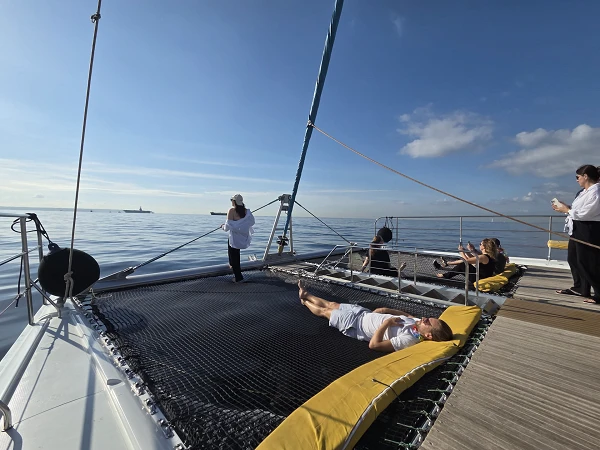 Personen entspannen auf einem Katamaran, während sie die ruhige Meereslandschaft und den blauen Himmel genießen. Eine Person liegt auf einem großen Netz vorne am Boot, während andere stehen und sitzen, um die Aussicht zu genießen.