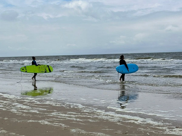 Zwei Surfer gehen mit ihren Surfbrettern, einem grünen und einem blauen, am Strand entlang ins Wasser, während die Wellen sanft an das Ufer rollen und der Himmel bewölkt ist.