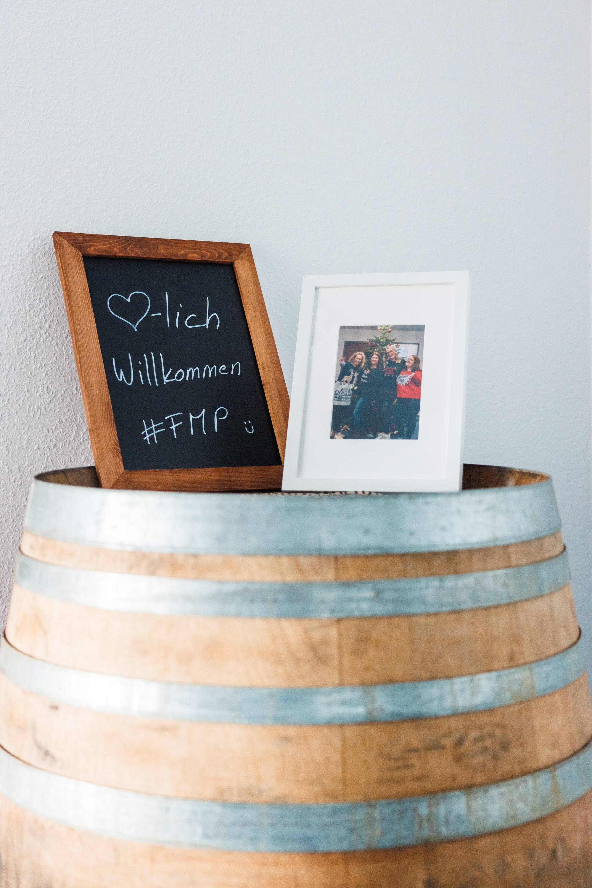Ein Holzfass dient als Ablage für ein kleines, gerahmtes Foto und eine Tafel mit der handgeschriebenen Begrüßung „Herzlich Willkommen #FMP“.