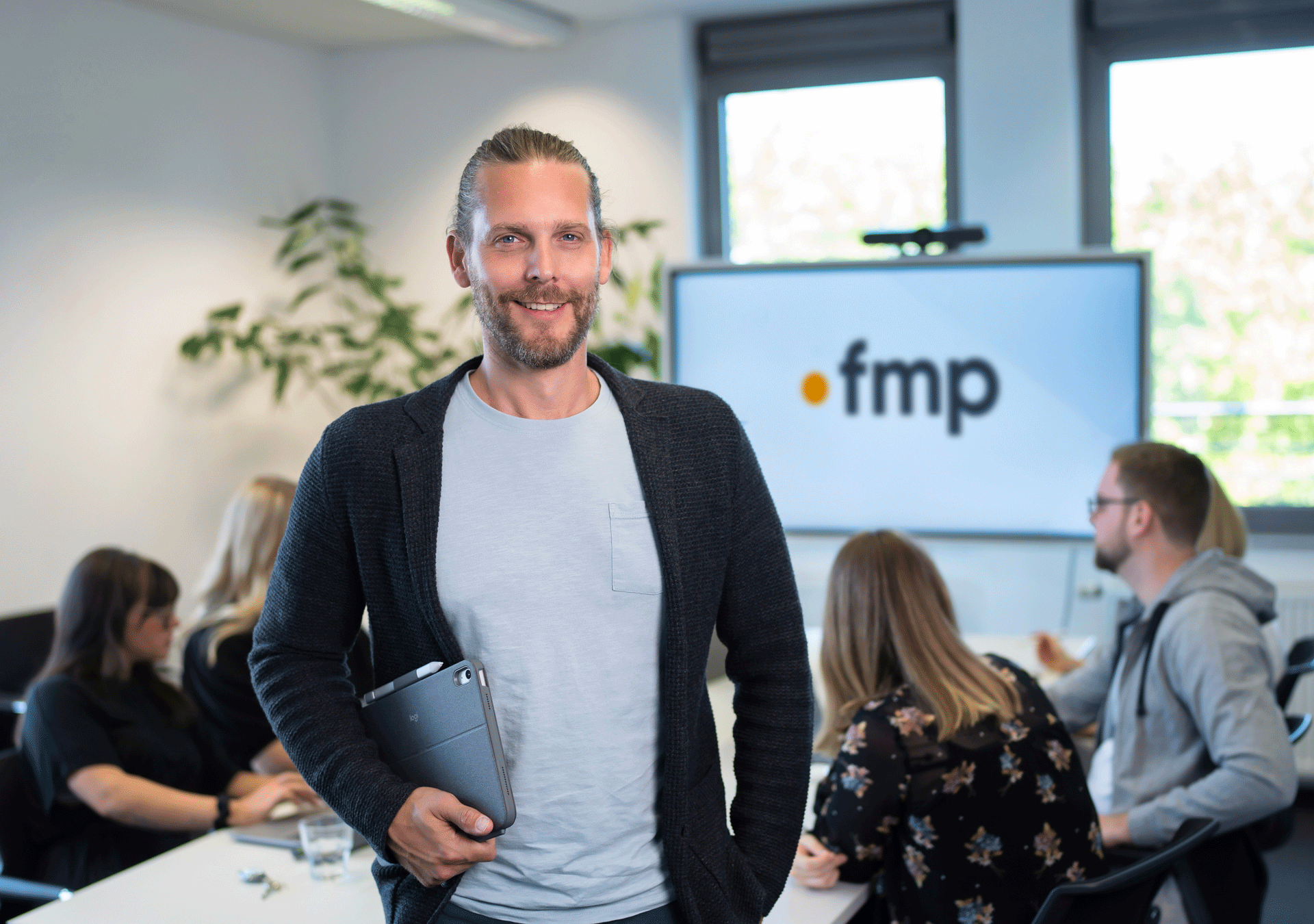 Ein Mann mit langen Haaren und einem breiten Lächeln steht in einem modernen Büro und hält ein Tablet in der Hand, während im Hintergrund eine Gruppe von Menschen an einem Tisch arbeitet und ein Bildschirm mit dem Logo „fmp“ zu sehen ist.