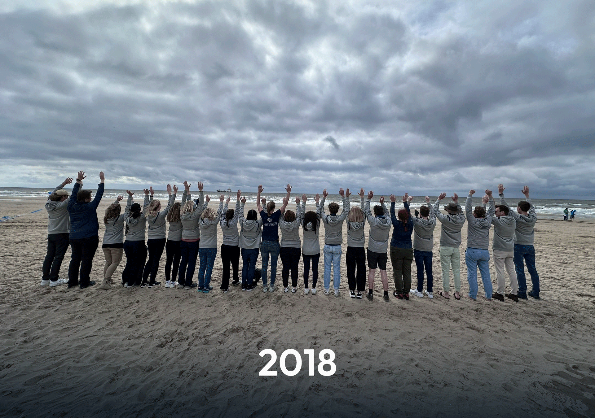 Eine Gruppe von Menschen steht am Strand, mit dem Gesicht zum Meer, und hebt beide Arme in die Luft. Die Personen tragen helle Sweatshirts und eine Wolkendecke ist am Himmel sichtbar. Unter dem Motiv steht die Jahreszahl "2018".
