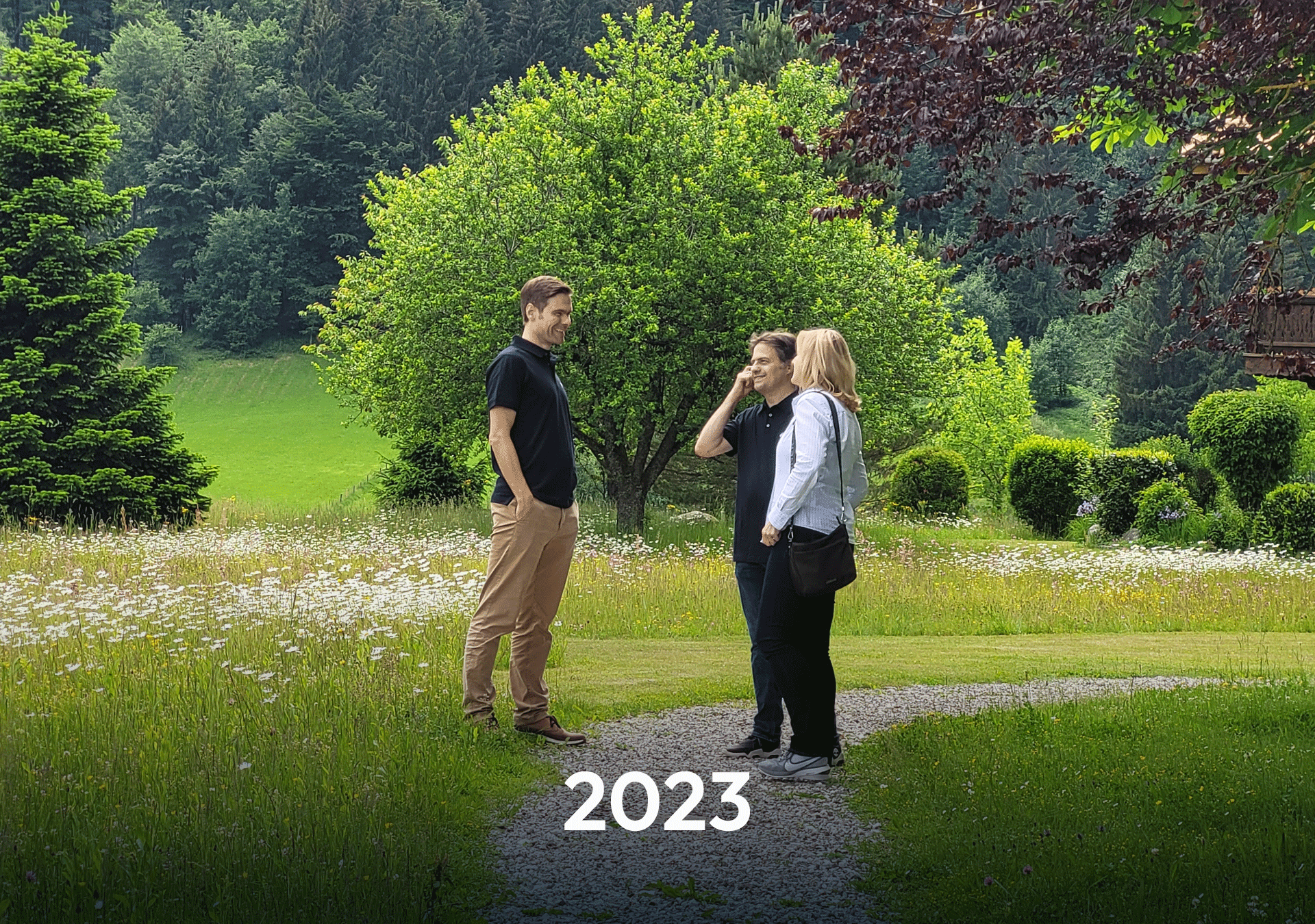 Drei Personen stehen auf einem Weg in einem grünen Garten, umgeben von Bäumen und Blumen, während sie miteinander sprechen. Im Vordergrund steht die Jahreszahl "2023".