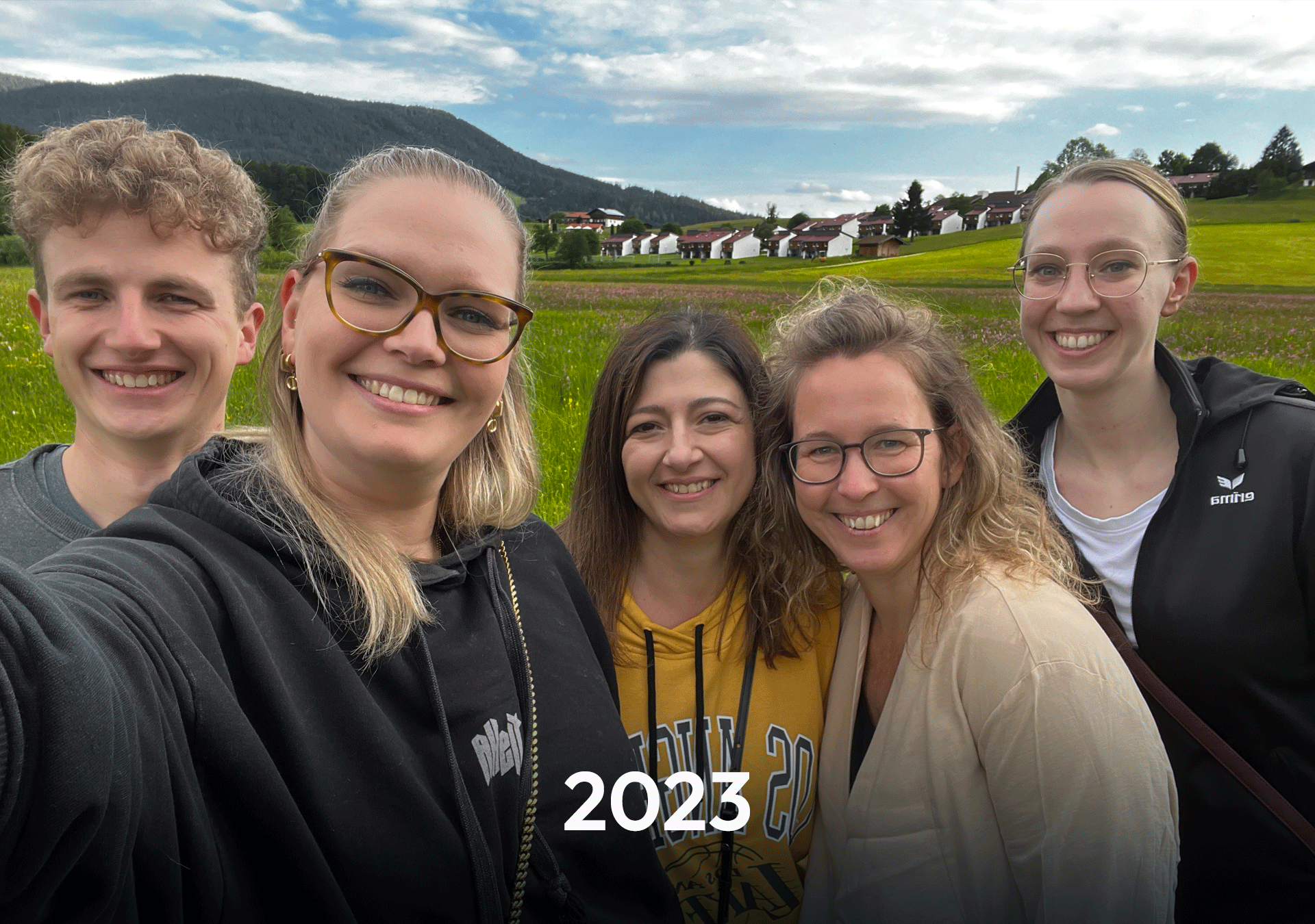 Eine Gruppe von fünf Personen macht ein Selfie vor einer grünen Wiesenlandschaft, umgeben von Bergen und einem Wohngebiet, mit dem Jahr „2023“ in der Mitte des Bildes.