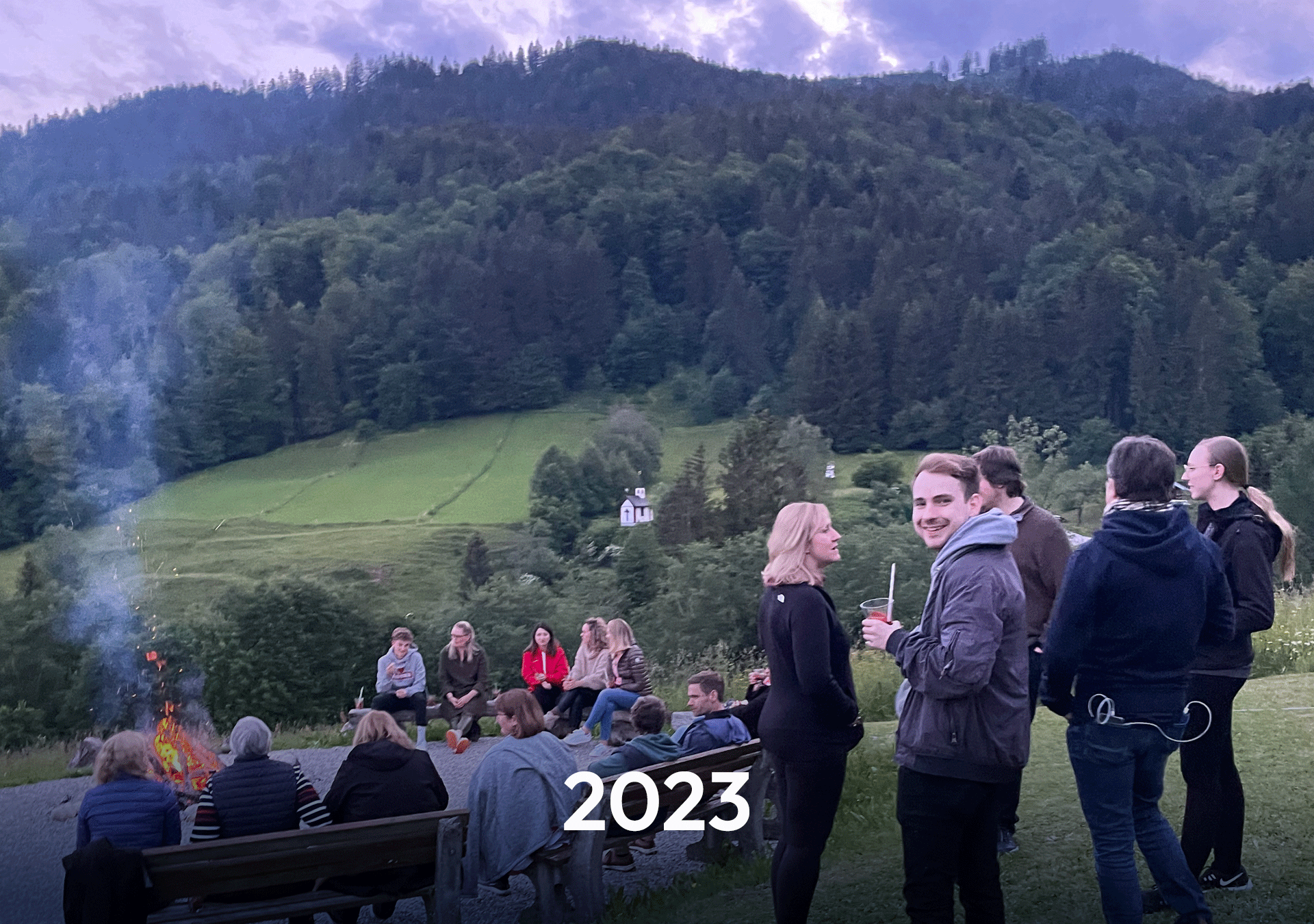Eine Gruppe von Menschen versammelt sich um ein Feuer in der Abenddämmerung, umgeben von einer malerischen Berglandschaft. Im Vordergrund steht ein Mann, der in die Kamera lächelt, während andere Personen mit Getränken plaudern. Im unteren Bereich des Bildes ist das Jahr „2023“ in großer Schrift zu sehen.