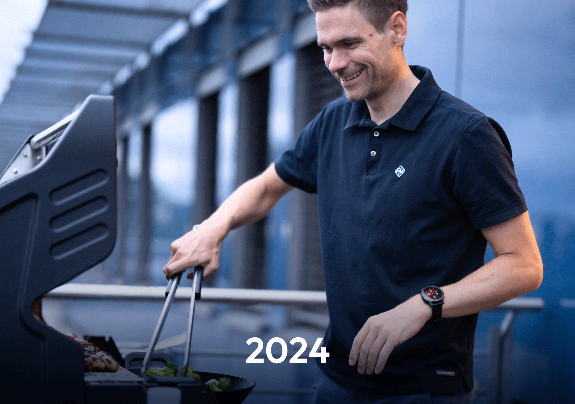 Ein Mann in einem schwarzen Polohemd steht an einem Grill und wendet mit Zangen Fleisch und Gemüse, während er lächelt. Im Vordergrund ist die Zahl „2024“ zu sehen.