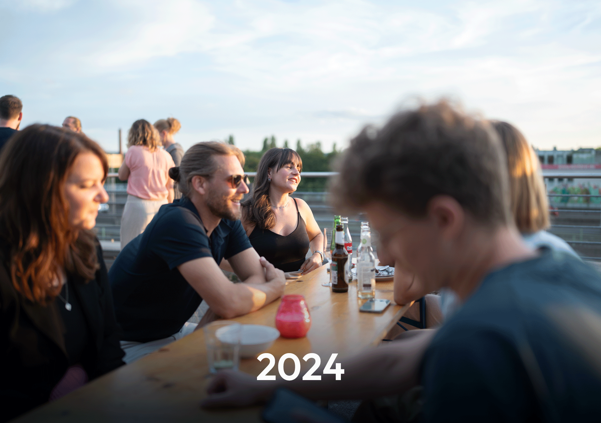 Eine Gruppe von Menschen sitzt an einem Holztisch auf einer Terrasse, während die Sonne untergeht. Einige Getränke stehen auf dem Tisch, und im Vordergrund ist das Jahr „2024“ in großen Zahlen abgebildet.