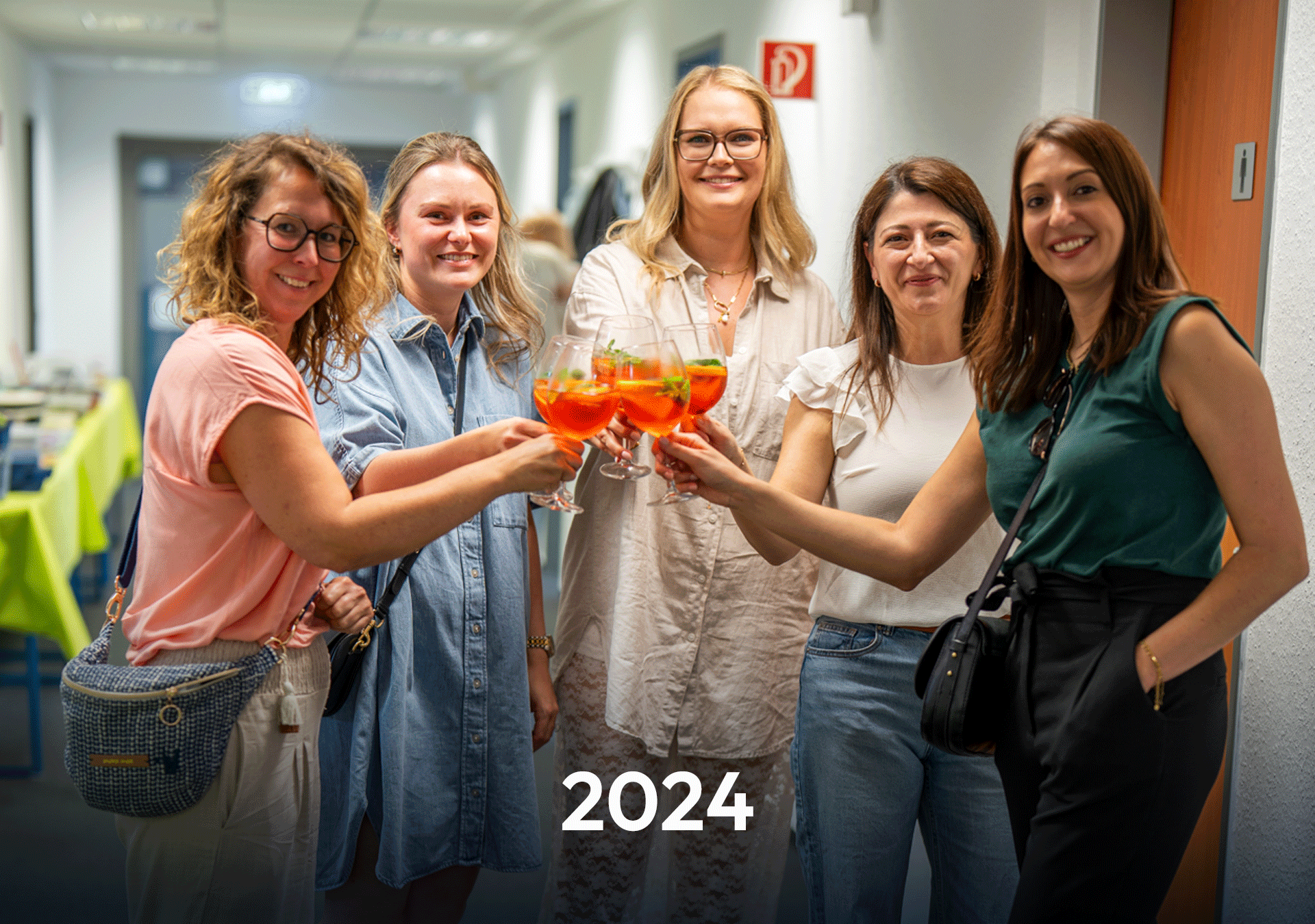 Eine Gruppe von fünf Frauen steht in einem Flur und hält Gläser mit bunten Cocktails in der Hand, während sie anstoßen. Im Hintergrund sind Teilbereiche eines Veranstaltungssaals sichtbar. Am unteren Bildrand steht die Zahl „2024“.