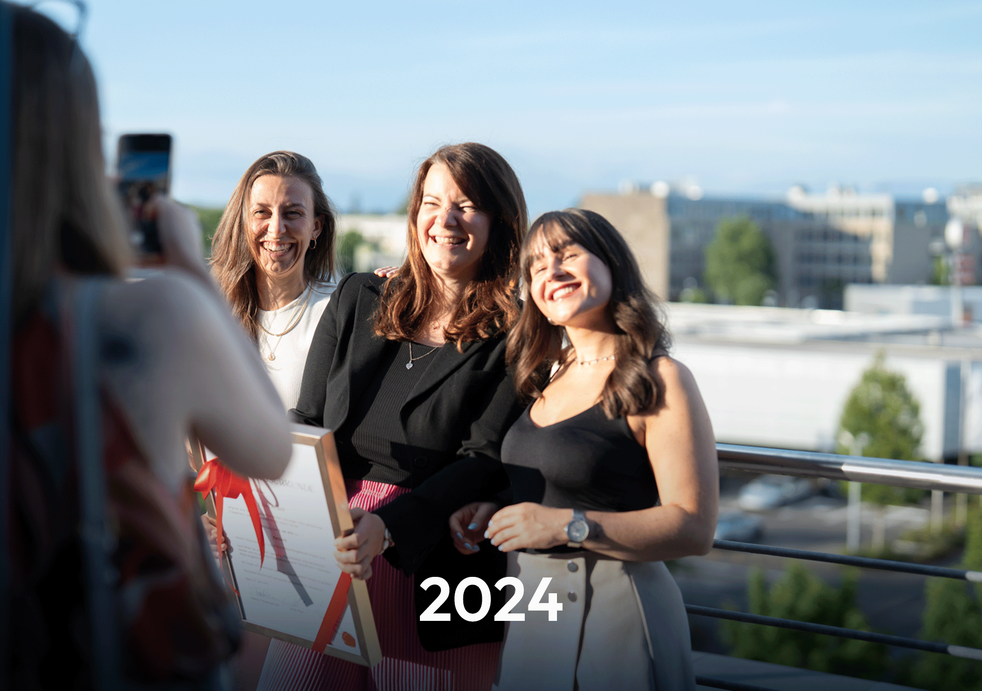 Drei Frauen posieren lächelnd für ein Foto auf einer Terrasse, während eine vierte Person sie mit einer Kamera aufnimmt. Im Vordergrund steht das Jahr „2024“ in großer Schrift.