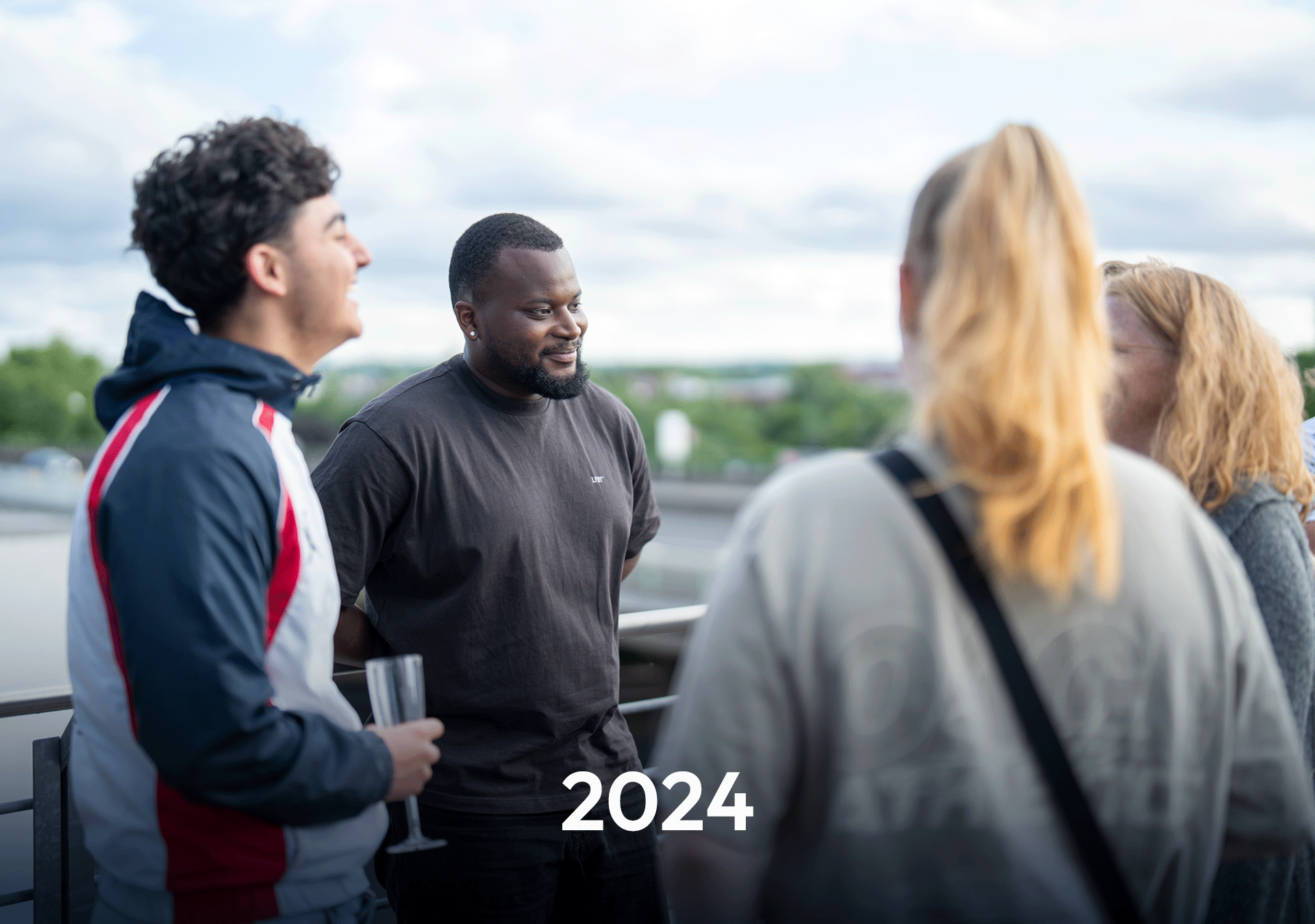 Eine Gruppe von vier Personen in casual Kleidung unterhält sich auf einem Balkon mit Blick auf eine grüne Landschaft und einen bewölkten Himmel. In der Mitte steht ein Mann mit einem freundlichen Lächeln. Der Text „2024“ steht prominent in der Mitte des Bildes.