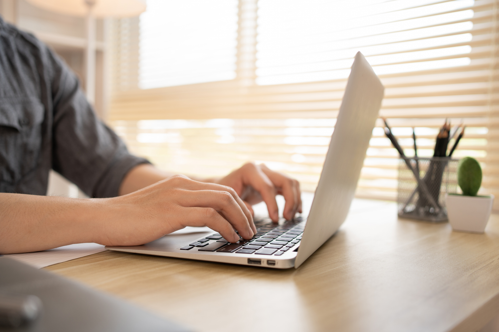 Eine Person tippt auf der Tastatur eines Laptops, während sie an einem Schreibtisch mit natürlichem Licht und horizontalen Jalousien arbeitet. Neben dem Laptop steht ein Behälter mit Stiften und eine kleine Pflanze.