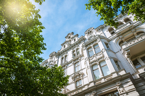 Die Fassade eines historischen Gebäudes mit dekorativen Elementen ist von grünen Baumblättern umgeben, während der Himmel blau und lichtdurchflutet ist.