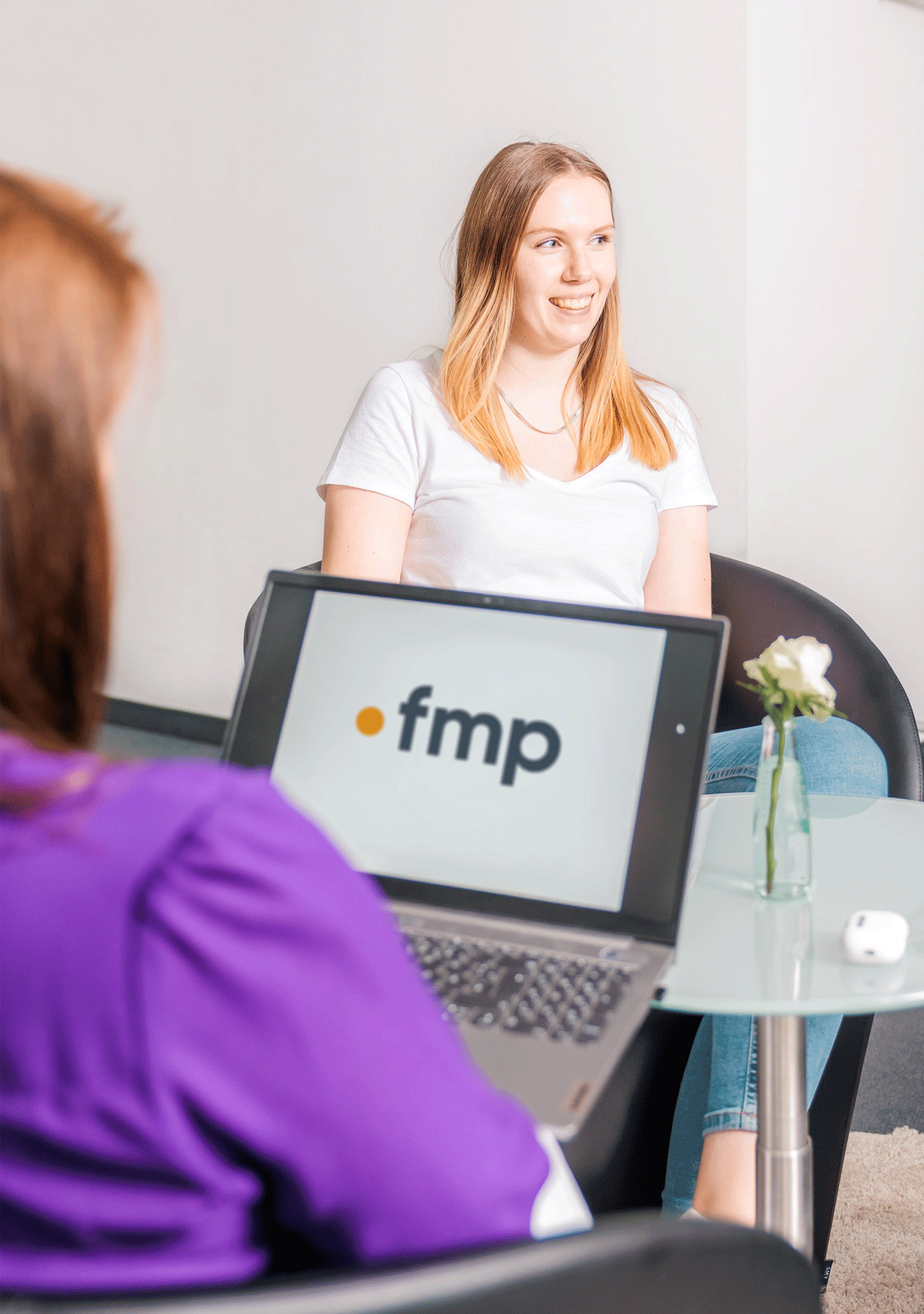 Eine Frau mit langen, blonden Haaren sitzt lächelnd auf einem Stuhl, während eine andere Person mit einer lila Bluse ihr gegenübersitzt. Auf einem Tisch steht ein Laptop mit dem Logo "fmp" und einer kleinen Vase mit einer Blume.