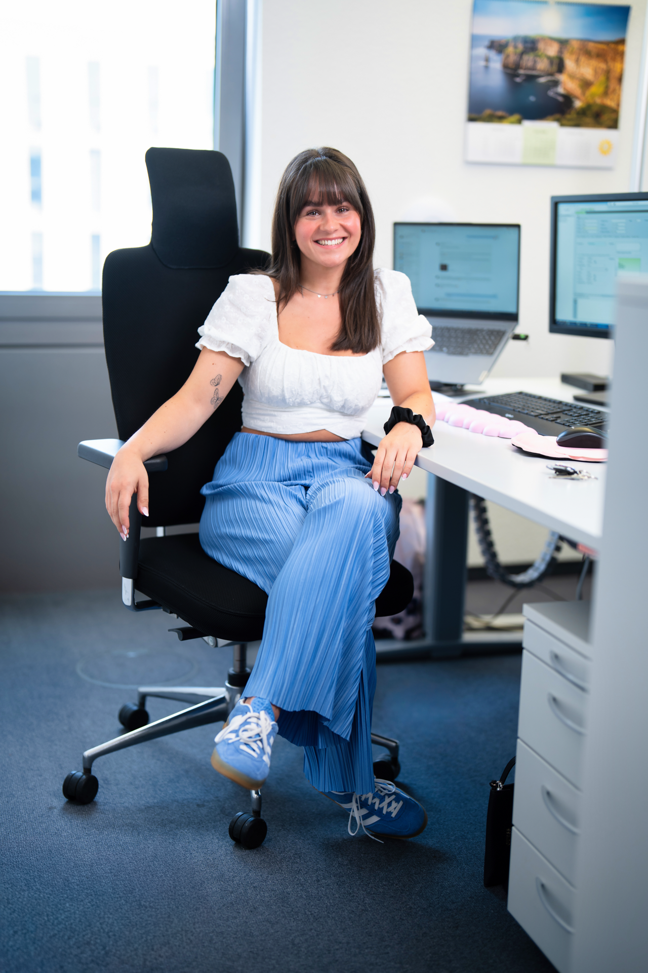 Eine lächelnde Frau mit langen, braunen Haaren sitzt entspannt auf einem Bürostuhl, trägt ein weißes Oberteil und hellblaue, gestreifte Hosen. Der Hintergrund zeigt einen modernen Arbeitsplatz mit zwei Computerbildschirmen und Büromaterialien.