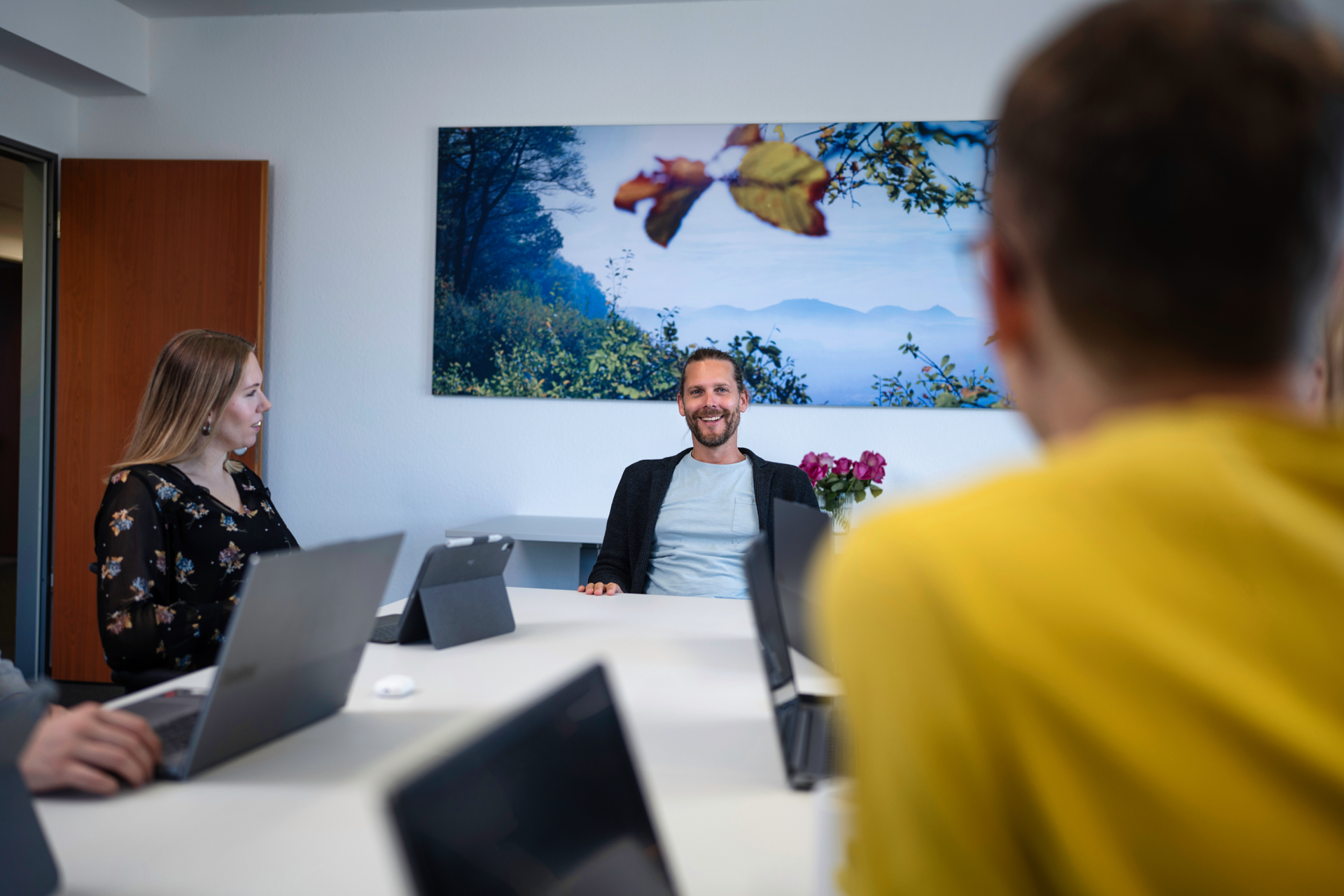 Eine Gruppe von fünf Personen sitzt an einem Tisch in einem Besprechungsraum; ein Mann in der Mitte lächelt, während er mit den anderen spricht. Im Hintergrund hängt ein Landschaftsbild mit Bäumen und Bergen.