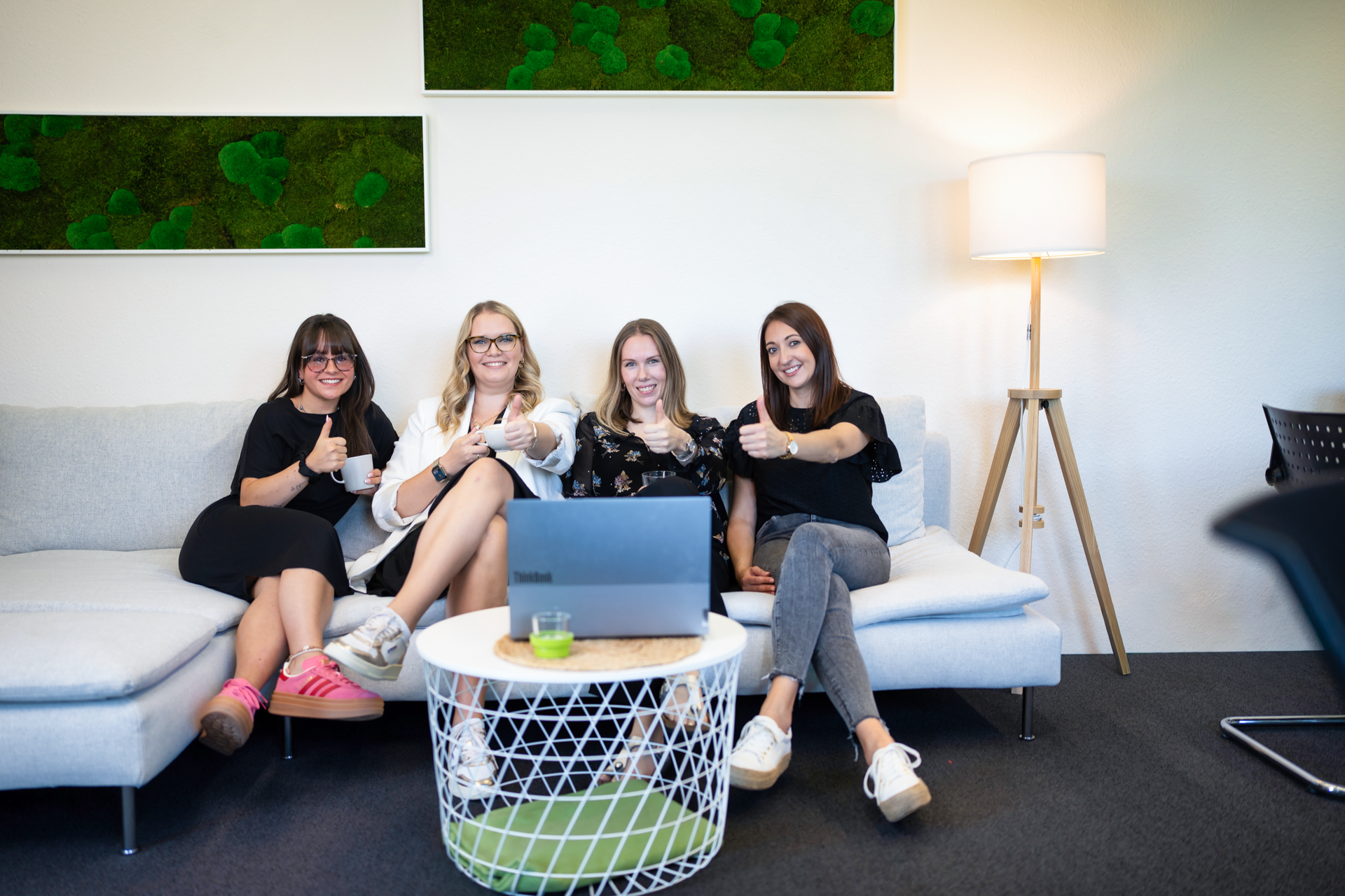 Vier Frauen sitzen auf einem hellen Sofa in einem modernen Büro. Sie zeigen Daumen nach oben und lächeln, während ein Laptop auf dem Tisch vor ihnen steht. Im Hintergrund sind grüne Wanddekorationen und eine Stehlampe zu sehen.