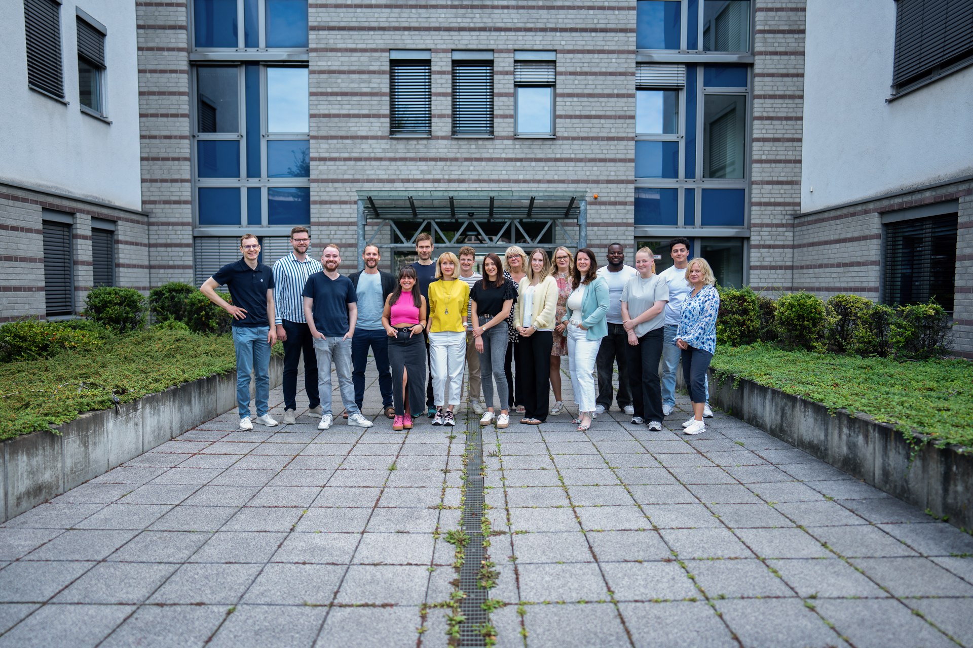 Eine Gruppe von 17 Personen steht lächelnd in einem Innenhof zwischen modernen Gebäuden, umgeben von grüner Bepflanzung und Steinen. Die Personen tragen verschiedene Freizeitoutfits und posieren freundlich für das Foto.