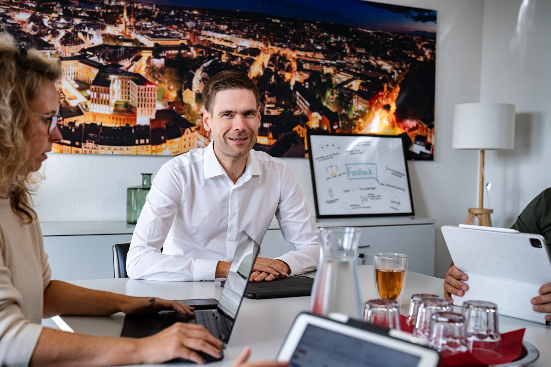 Ein Mann in einem weißen Hemd sitzt an einem Tisch in einem Besprechungsraum und lächelt, während um ihn herum mehrere Personen mit Laptops und einem Tablet beschäftigt sind. Im Hintergrund ist ein beleuchtetes Stadtbild an der Wand zu sehen.