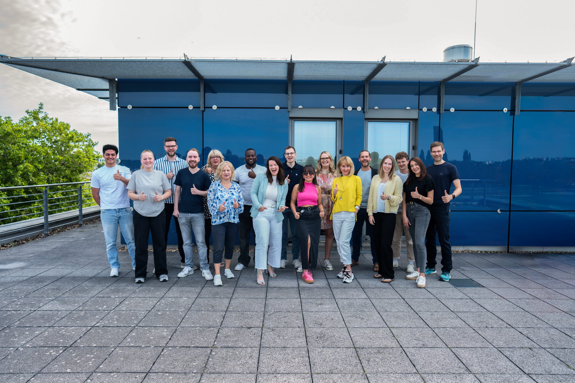 Eine Gruppe von 20 Personen steht lächelnd auf einem gepflasterten Bereich vor einem modernen, blauen Gebäude. Die Anwesenden machen Daumen-hoch-Gesten und tragen unterschiedliche, lässige Kleidung. Im Hintergrund sind Bäume und Fenster des Gebäudes sichtbar.