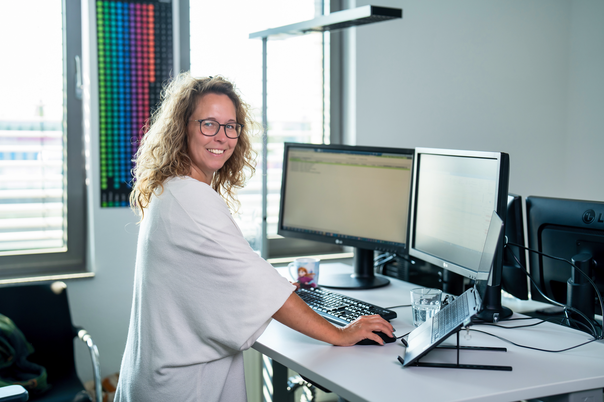 Eine lächelnde Frau mit lockigem Haar sitzt an einem Schreibtisch und arbeitet an zwei Monitoren, während sie auf die Kamera blickt. Die Umgebung wirkt modern und hell, mit einer Glaswasserflasche und einem Ständer für den Laptop auf dem Tisch.