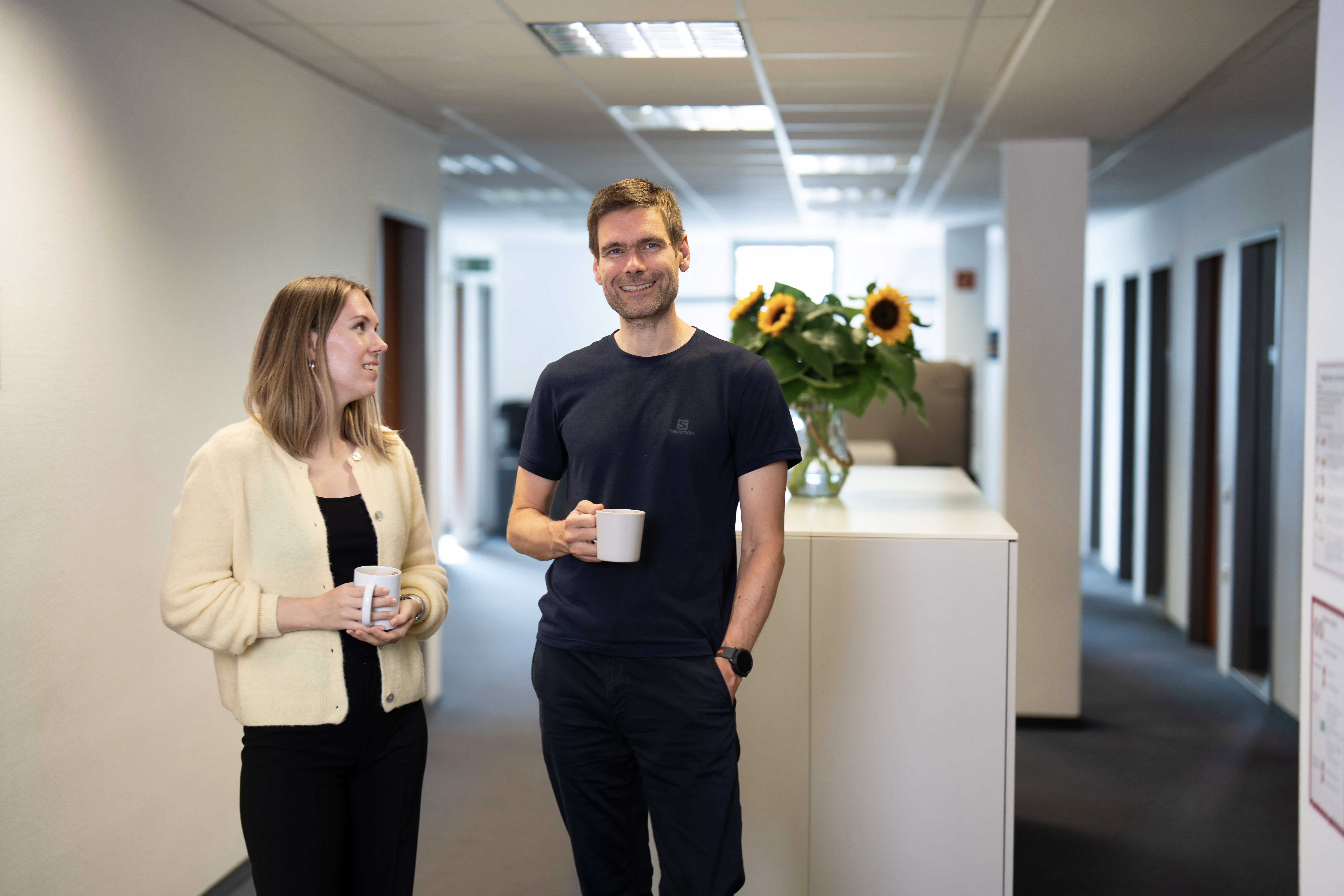 Ein Mann und eine Frau stehen in einem modernen Büroflur, beide halten eine Tasse in der Hand. Die Frau schaut den Mann an und lächelt, während im Hintergrund ein großer Blumenstrauß mit Sonnenblumen zu sehen ist.