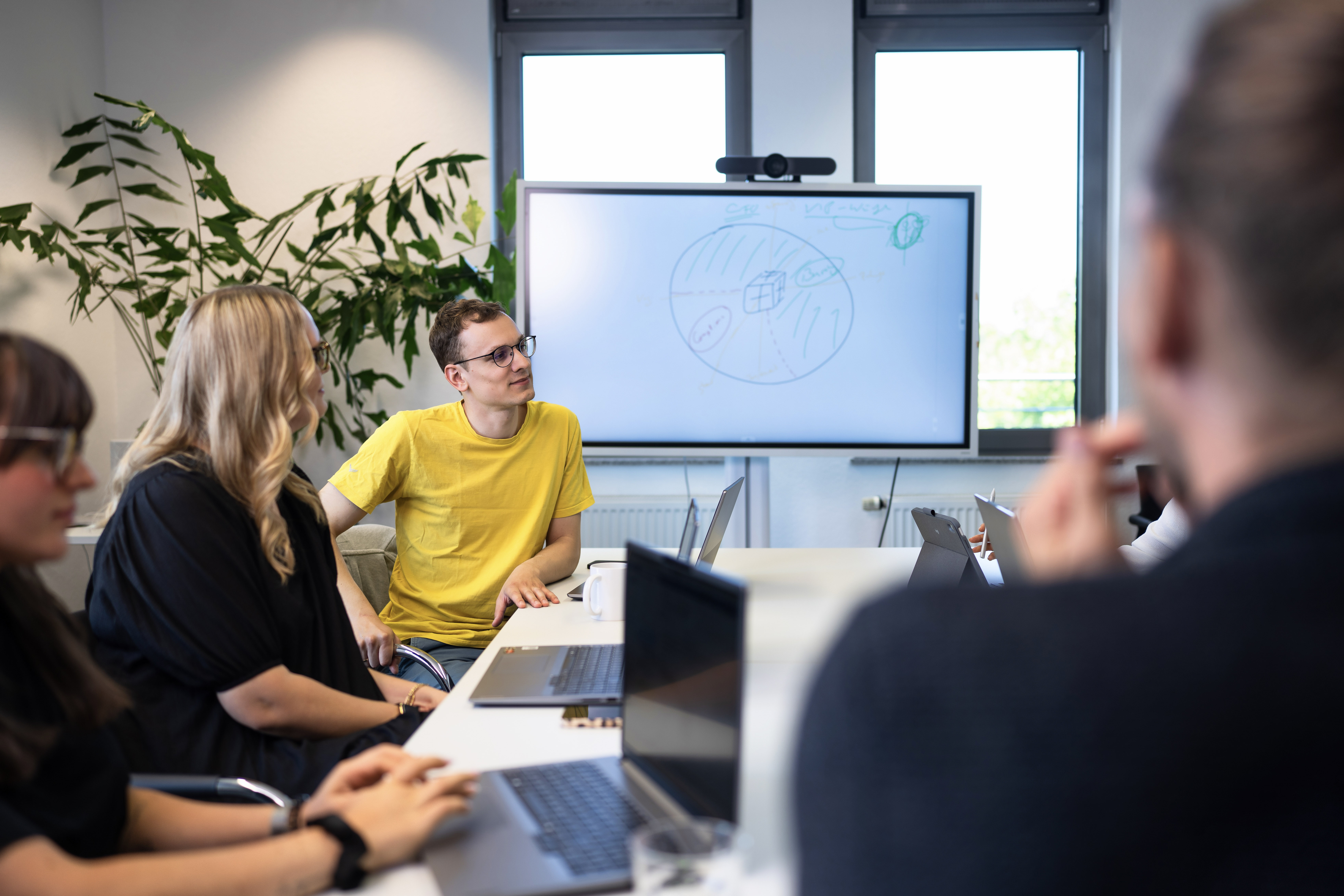 Eine Gruppe von Menschen sitzt an einem Konferenztisch, während ein junger Mann in gelbem T-Shirt auf einen Bildschirm zeigt, auf dem eine Zeichnung zu sehen ist. Im Hintergrund sind Pflanzen und Fenster erkennbar.