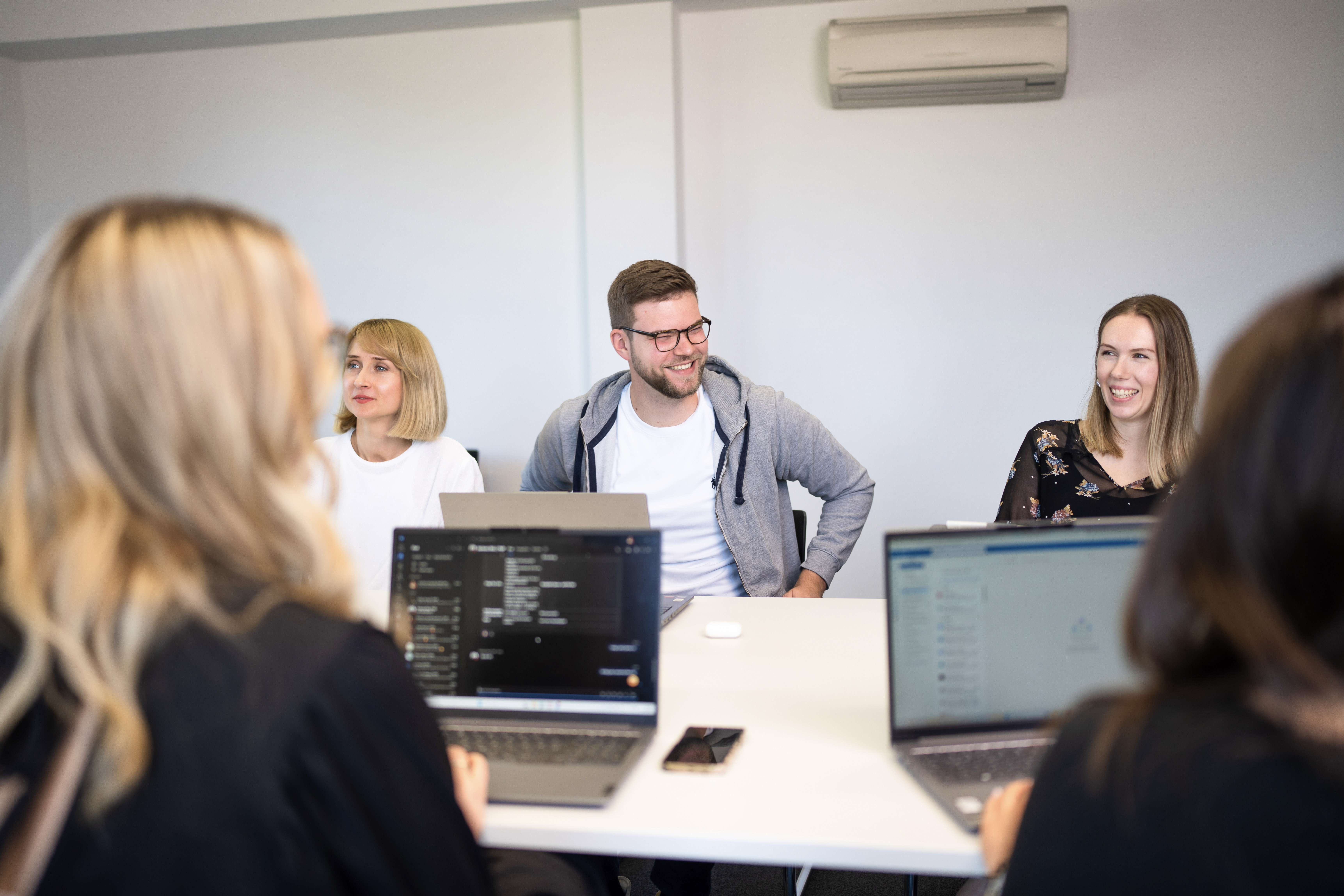 Eine Gruppe von vier Personen sitzt an einem Tisch in einem modernen Büro, während sie sich unterhalten. Zwei Laptops sind auf dem Tisch sichtbar, und der Mann in der Mitte trägt eine Brille sowie ein graues Sweatshirt.