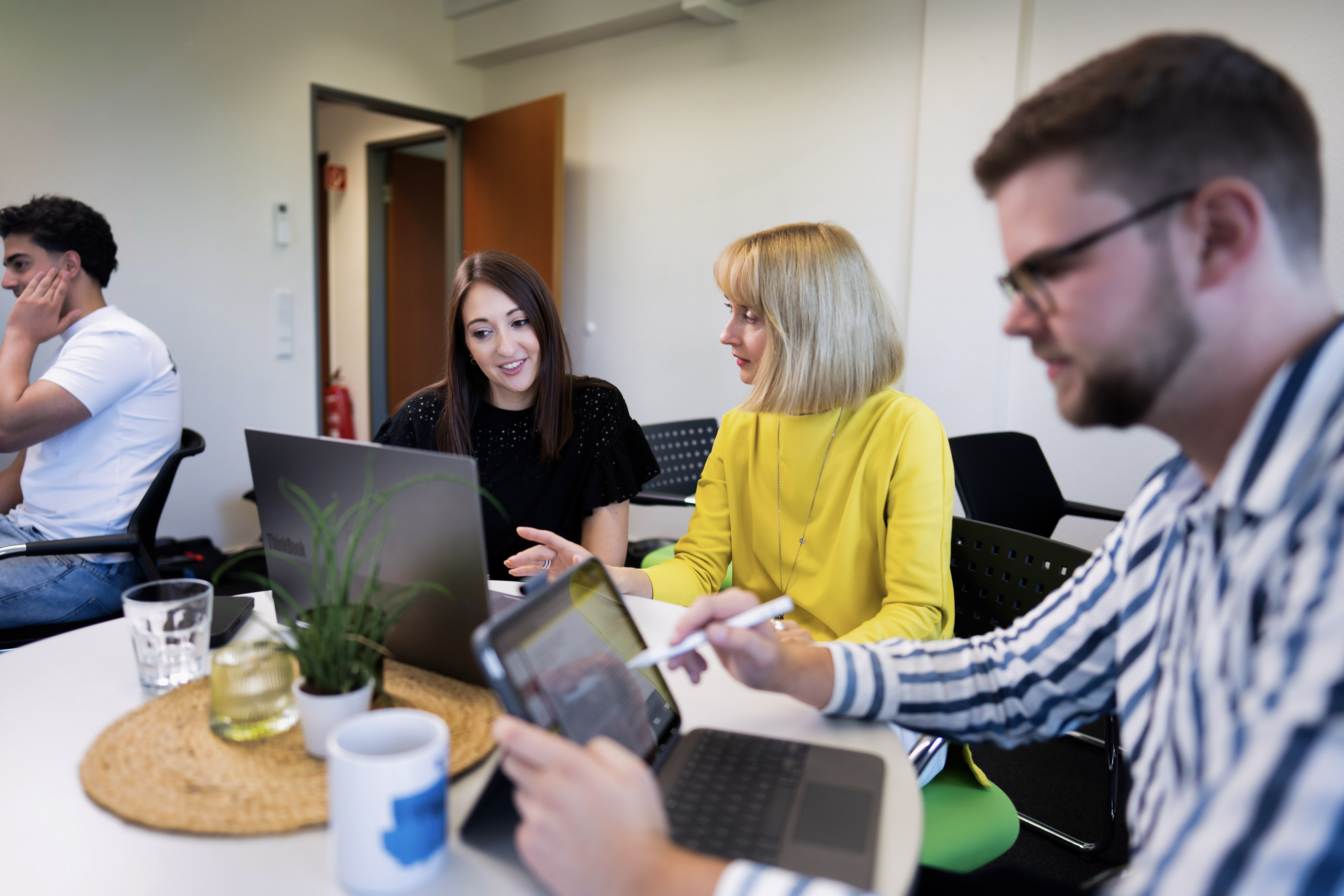 Zwei Personen am Tisch sehen sich eine Präsentation auf einem Laptop an, während eine dritte Person im Hintergrund mit einem Smartphone beschäftigt ist. Auf dem Tisch stehen Getränke und eine kleine Pflanze.