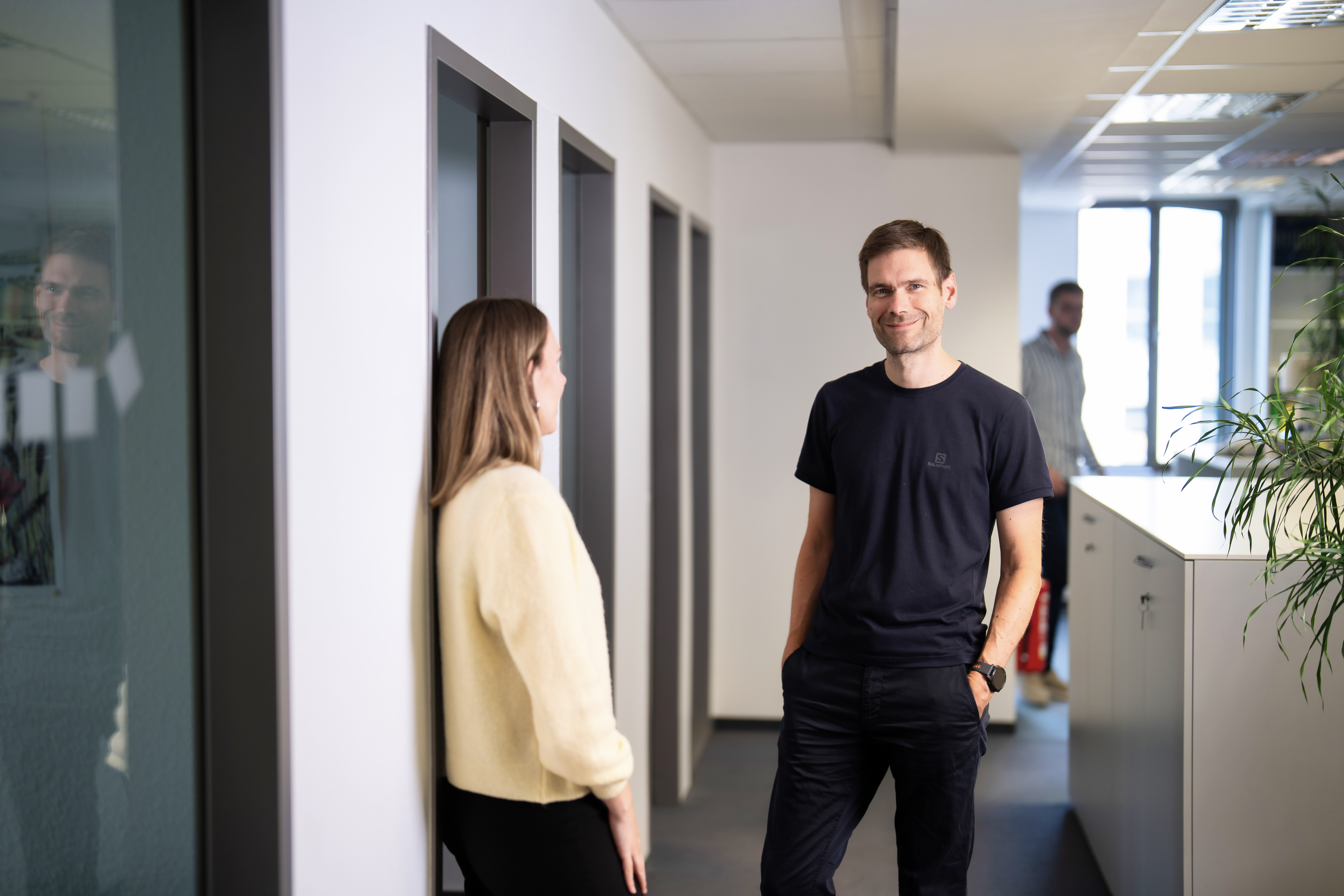 Zwei Personen stehen in einem modernen Büroraum, während sie sich unterhalten; im Hintergrund sind weitere Büros und ein Pflanzenarrangement sichtbar.