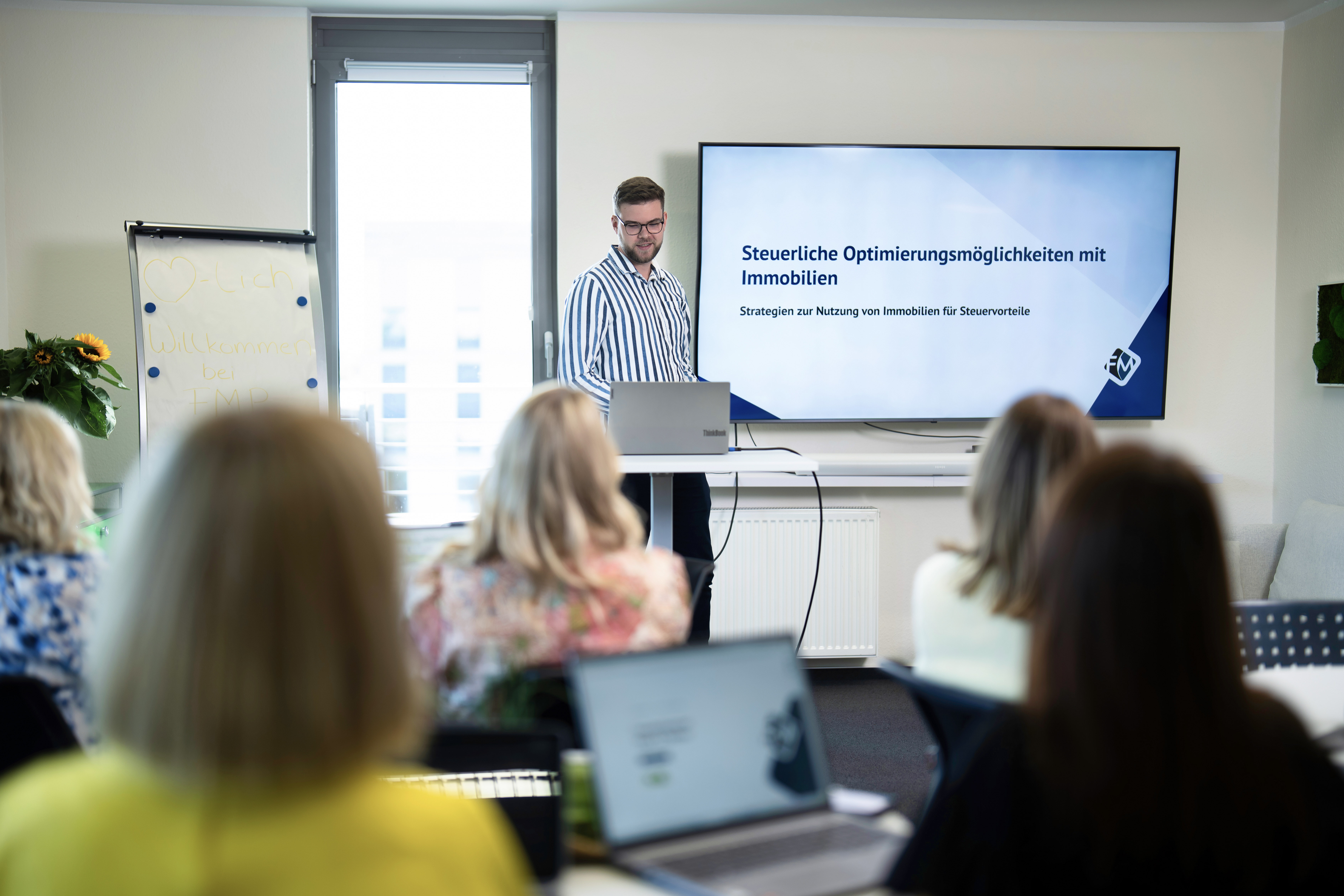 Ein Mann präsentiert vor einer Gruppe von Zuhörern in einem Seminarraum, während im Hintergrund eine Bildschirmpräsentation mit dem Titel „Steuerliche Optimierungsmöglichkeiten mit Immobilien“ zu sehen ist. Der Mann spricht über Strategien zur Nutzung von Immobilien für Steuerzwecke. Die Zuhörer sind teilweise mit Laptops beschäftigt.
