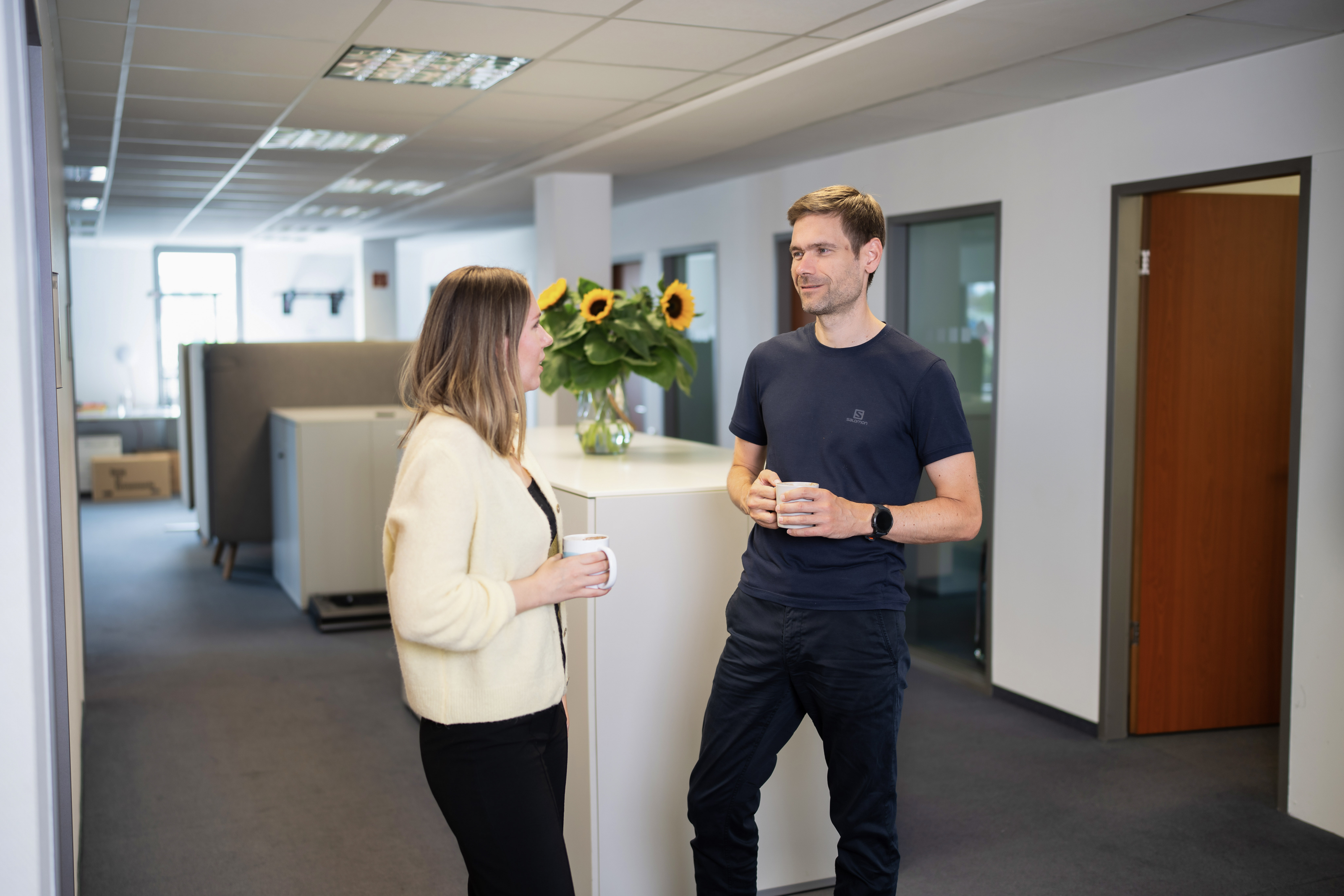 Zwei Personen unterhalten sich in einem modernen Büroflur, während sie dampfende Becher in den Händen halten. Im Hintergrund sind Fenster und ein Strauß Sonnenblumen zu sehen.